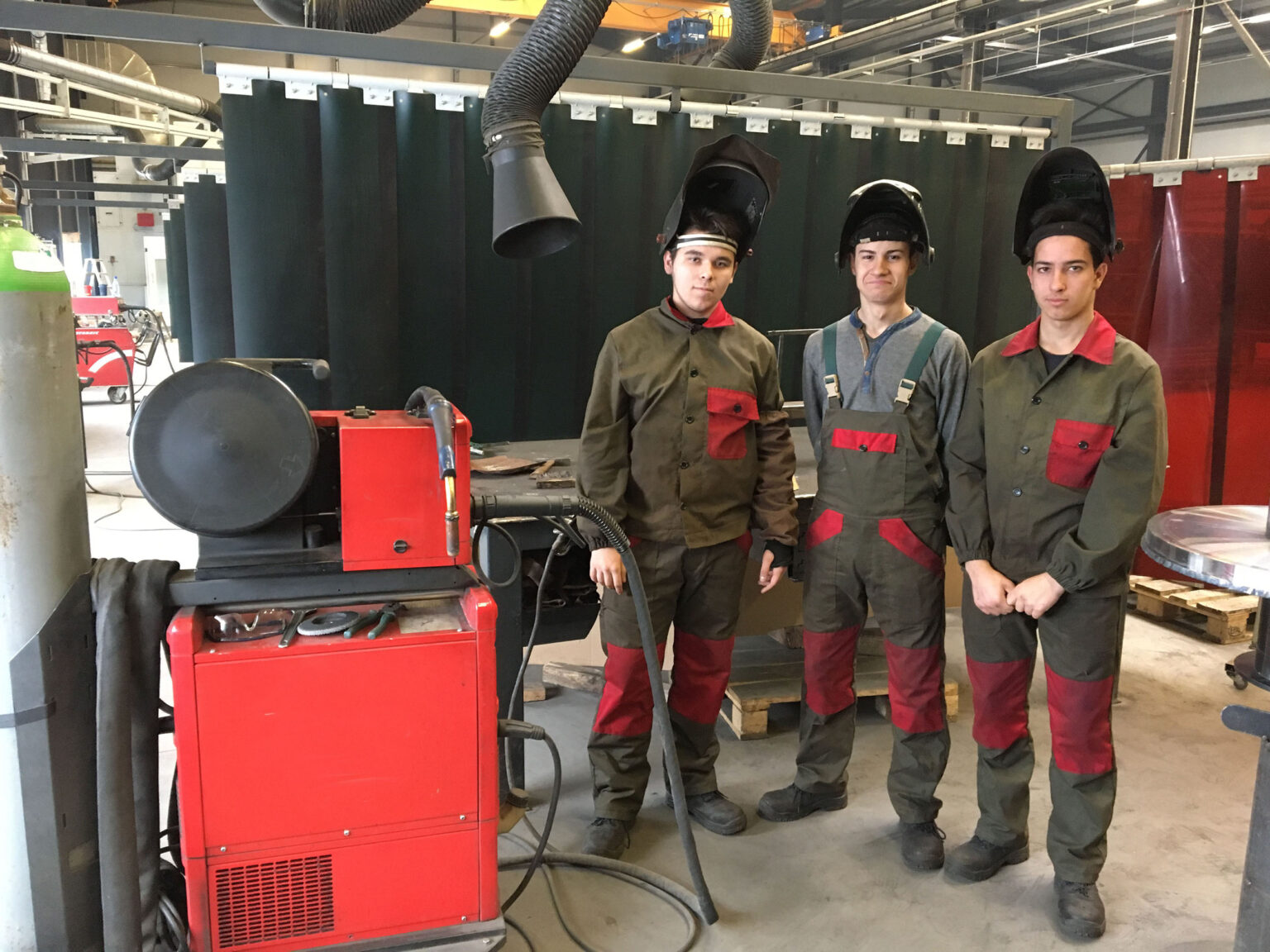Tres trabajadores, vestidos con ropa de protección, se encuentran junto a una máquina de soldadura en un taller.