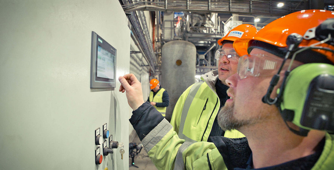 Dos trabajadores con ropa de protección manejan un panel de control en una planta industrial.