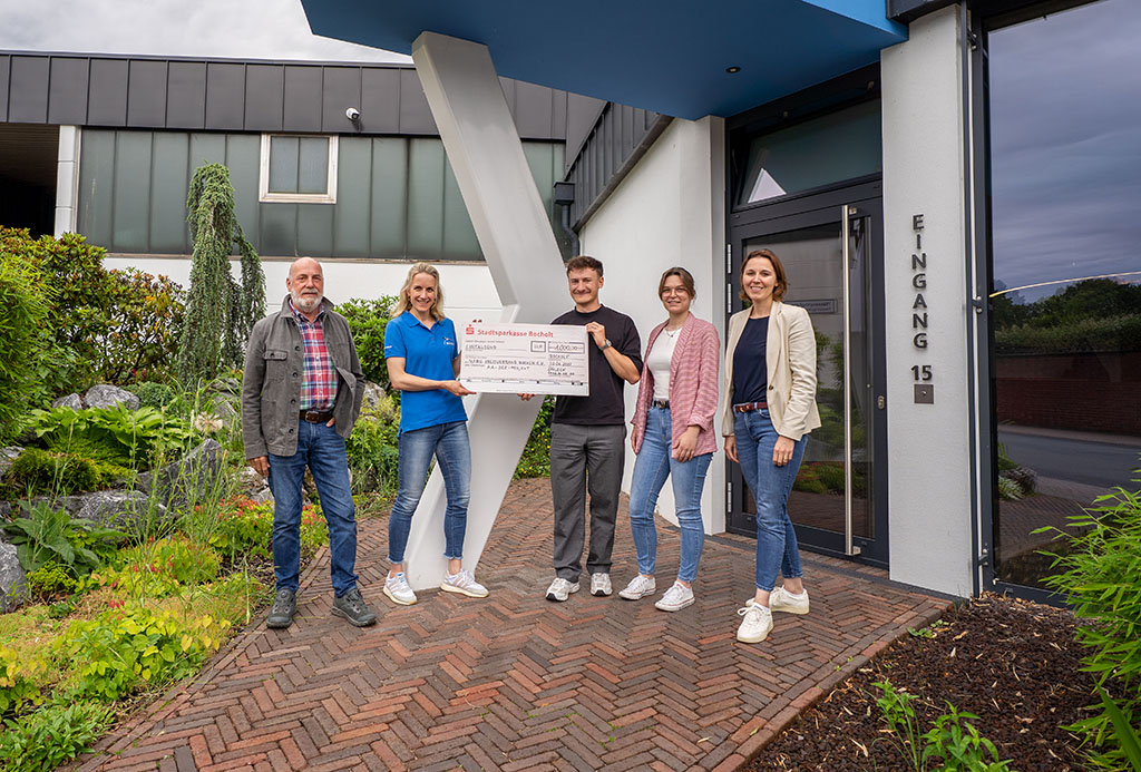 Un grupo de cinco personas sostiene un cheque de gran tamaño frente a un edificio.