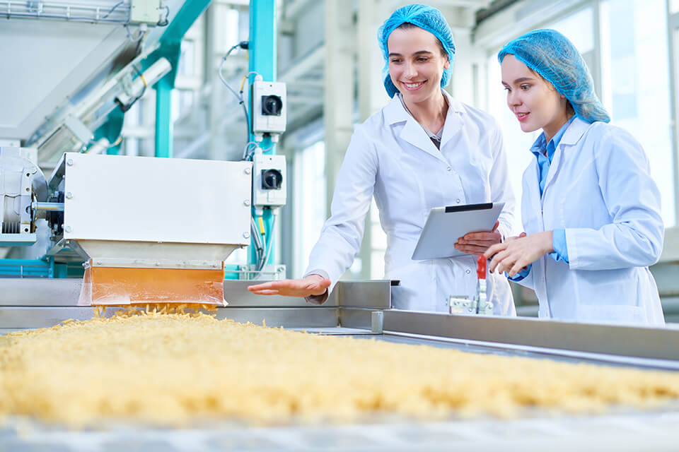Dos mujeres con batas de laboratorio inspeccionan una línea de producción de material a granel.